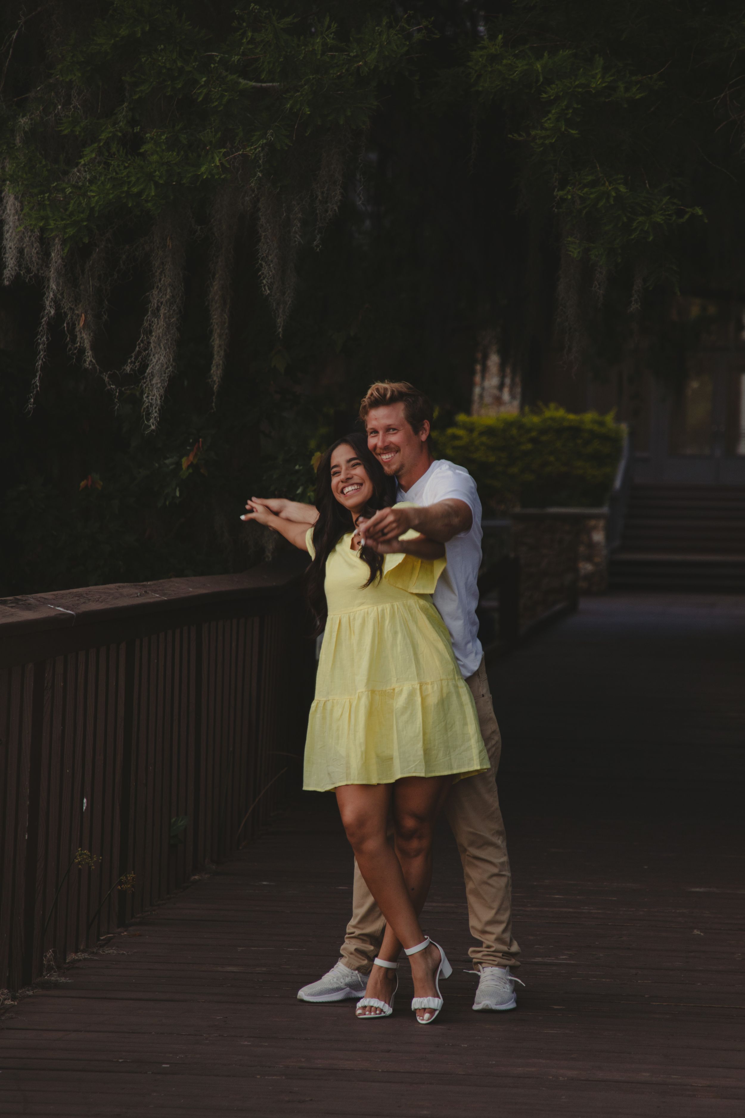 A couple in a yellow dress and khaki pants share a romantic moment on a wooden boardwalk at dusk.