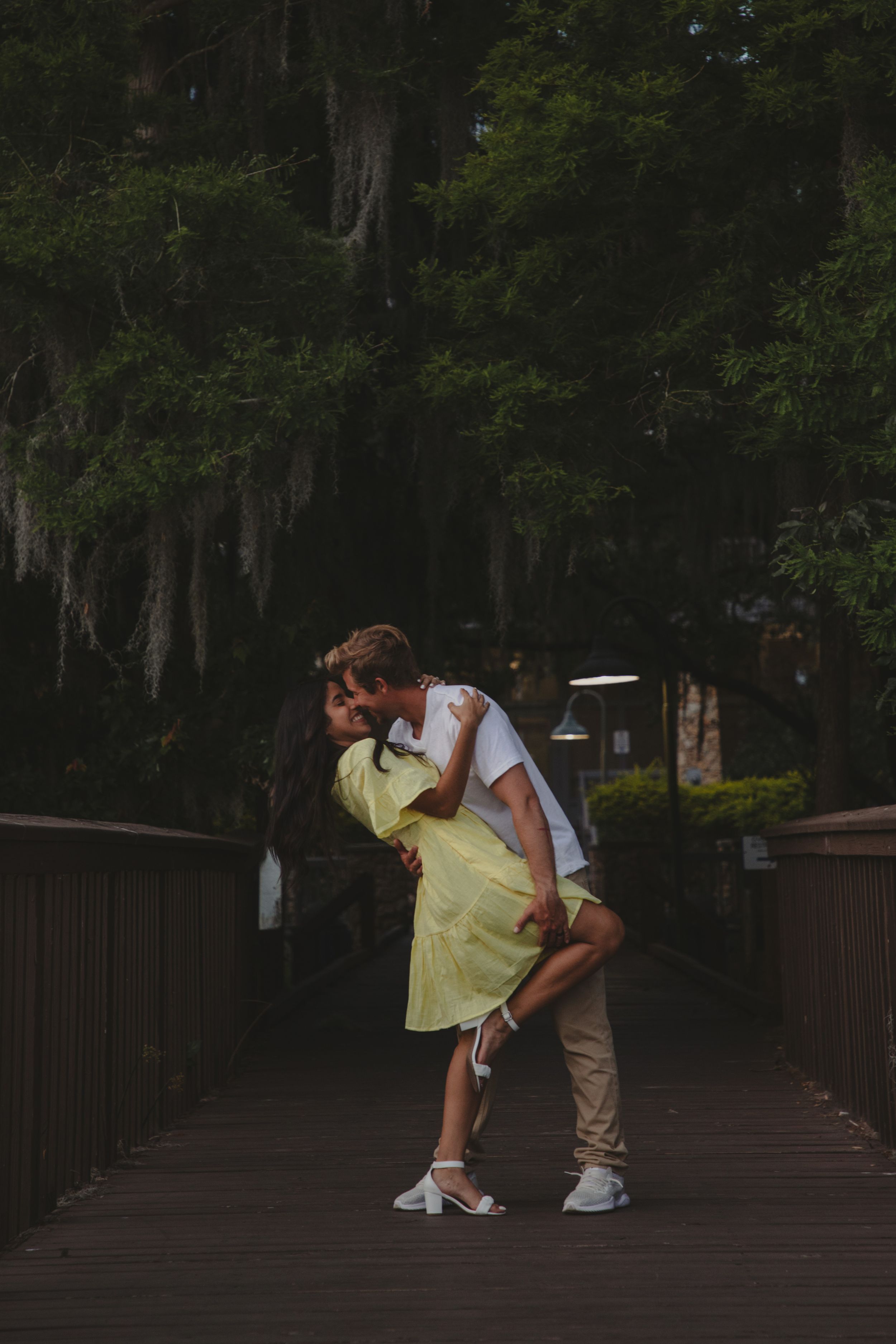 Two people share an intimate moment on a wooden boardwalk surrounded by Spanish moss draped trees at dusk.