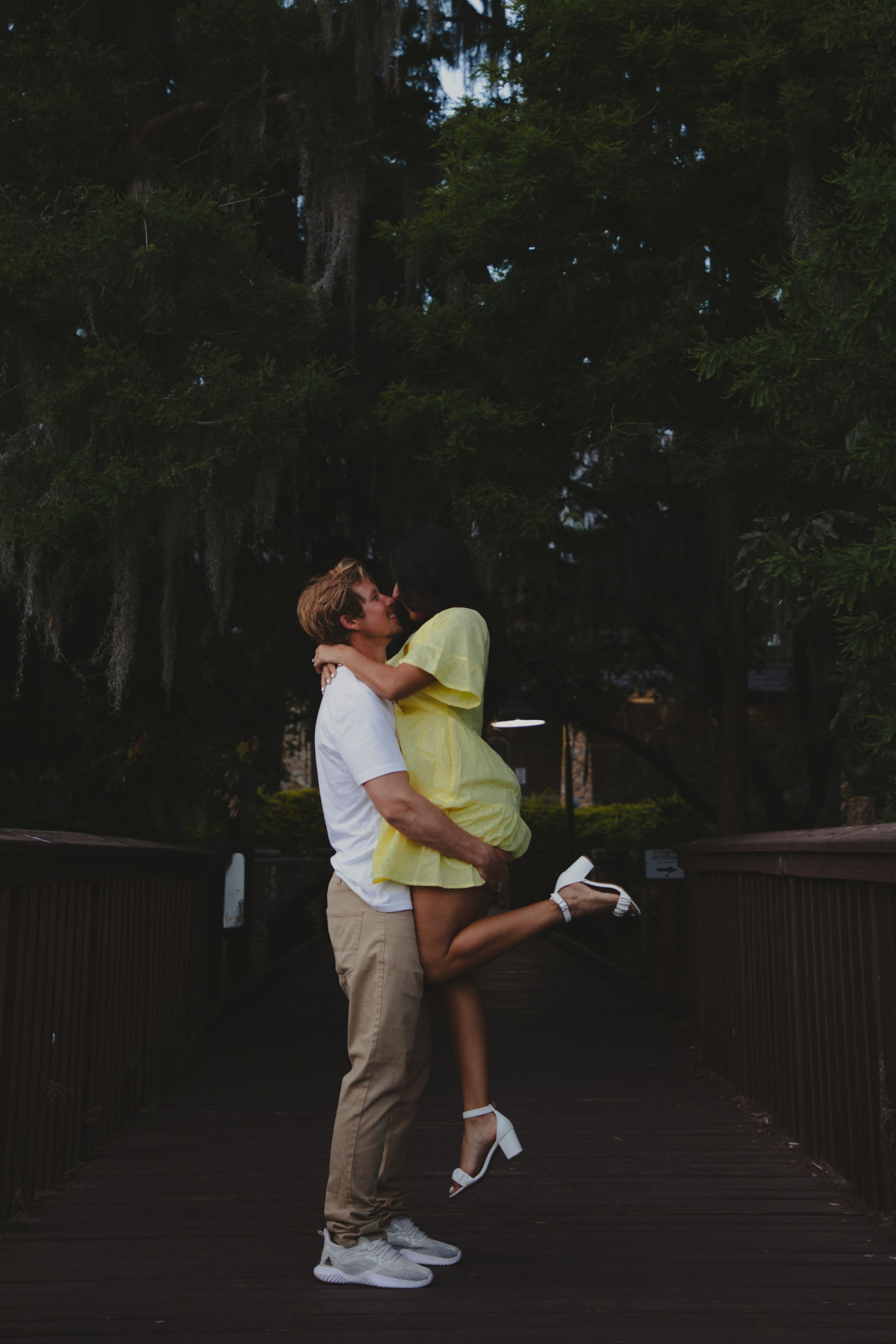 A romantic couple embraces and kisses on a wooden bridge surrounded by lush trees at dusk.