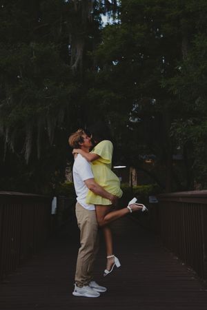 A romantic couple embraces and kisses on a wooden bridge surrounded by lush trees at dusk.