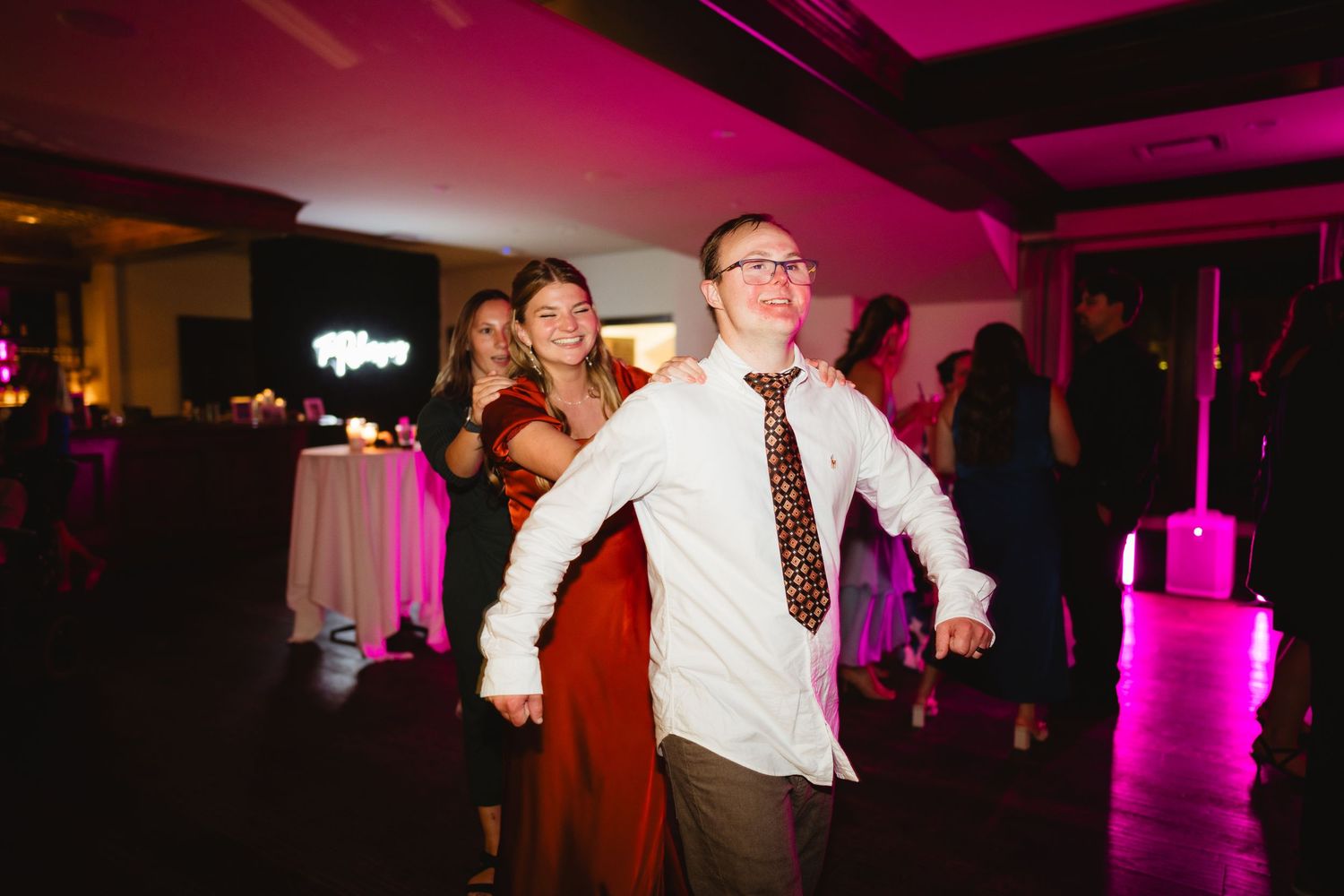 People dancing and celebrating at a wedding reception with pink and purple lighting illuminating the dance floor.