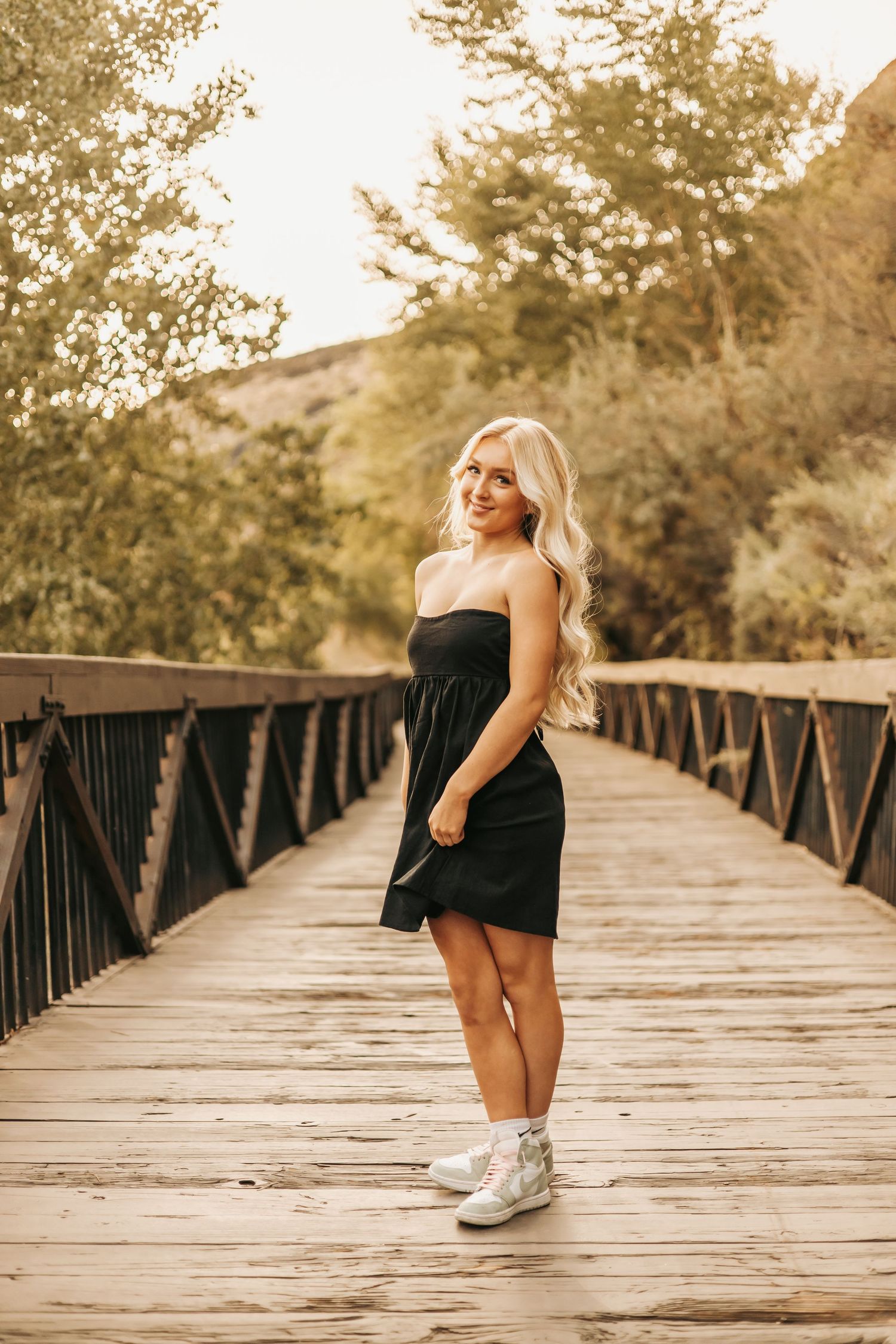 Woman in black strapless dress and white sneakers poses on wooden bridge during golden hour sunset.