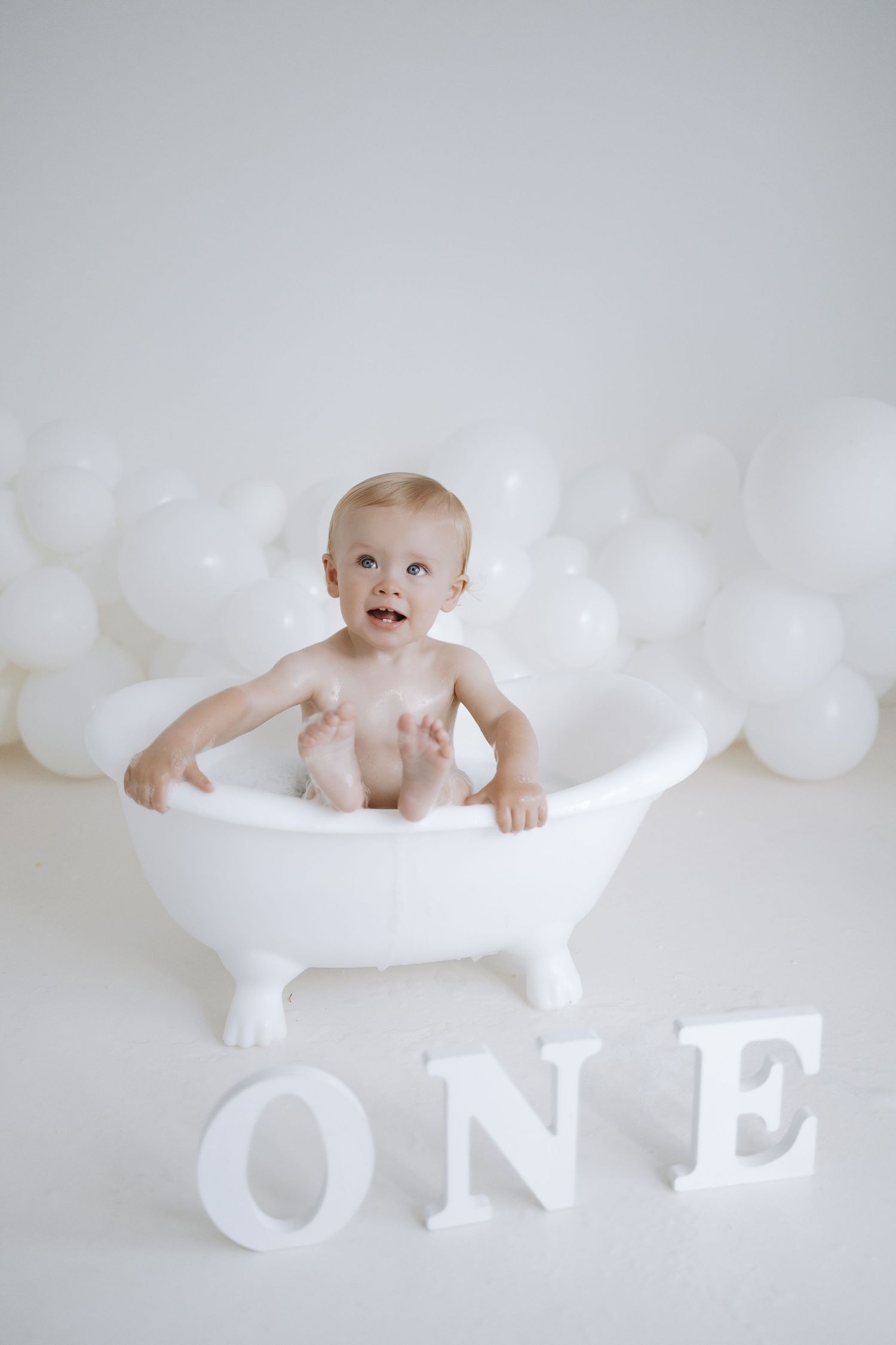 A baby in white surroundings poses with large decorative letters spelling out 'ONE' for their first birthday milestone photo.