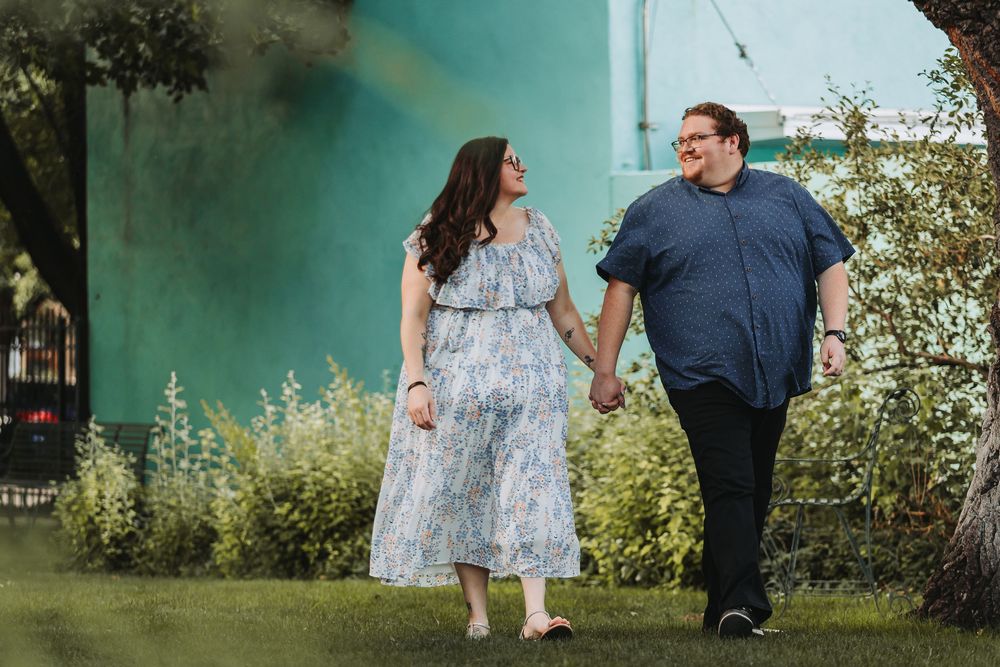 A couple in casual attire walks together through a lush garden setting with a turquoise wall in the background.