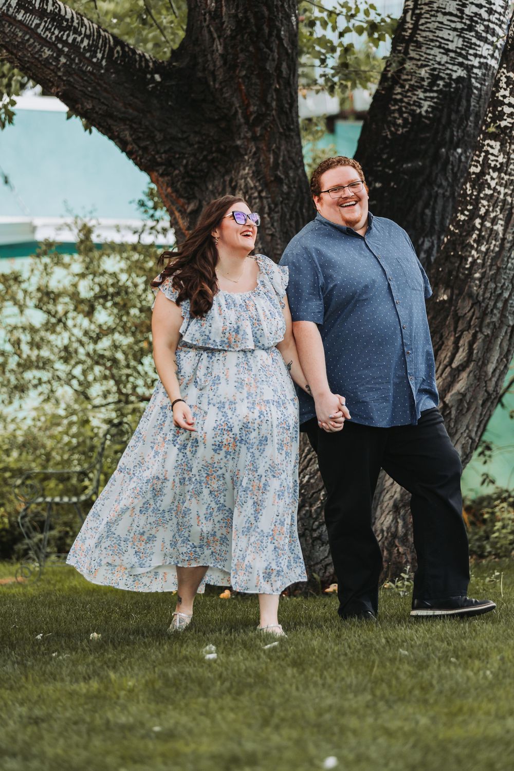 A couple in casual attire shares a playful moment under a tree on a sunny day in a park setting.