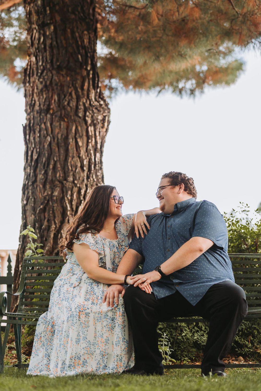 A romantic couple sits together on a bench under a large pine tree, sharing a tender moment in a garden setting.