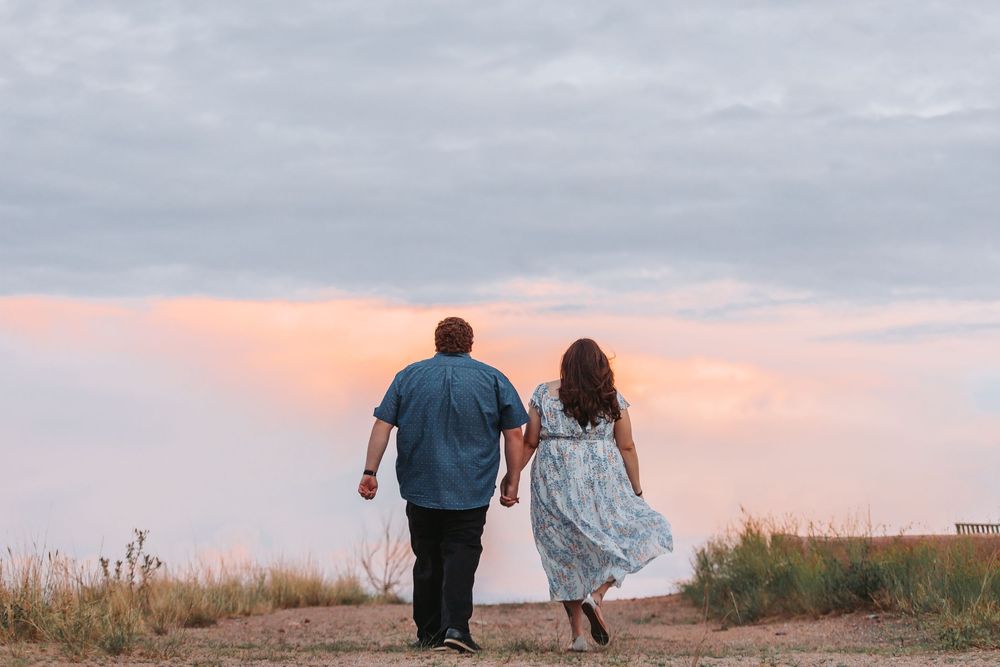 A couple stands together watching a beautiful pink and orange sunset over a grassy field.