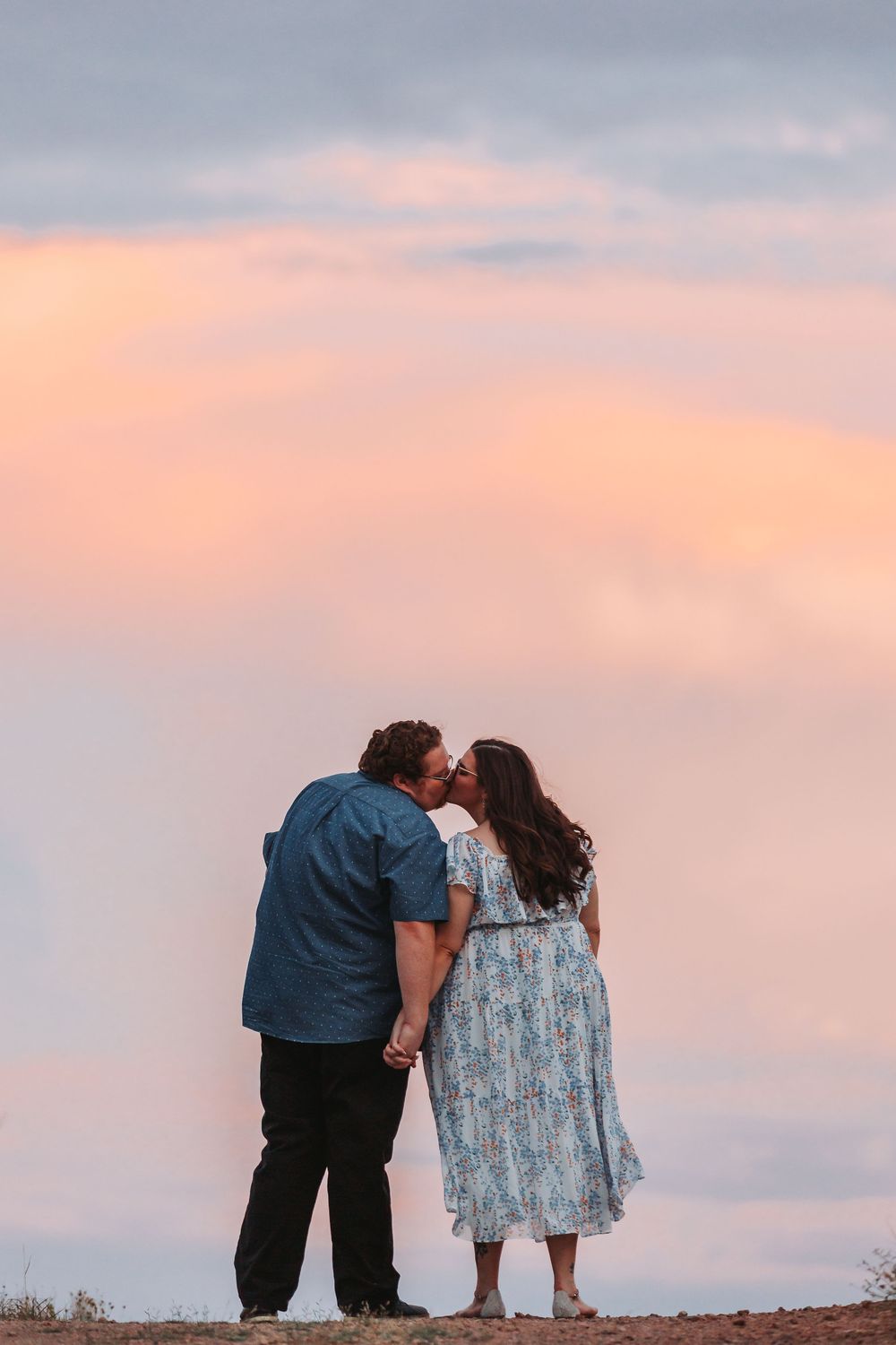 A couple shares a tender moment against a pastel sunset sky on a hilltop.