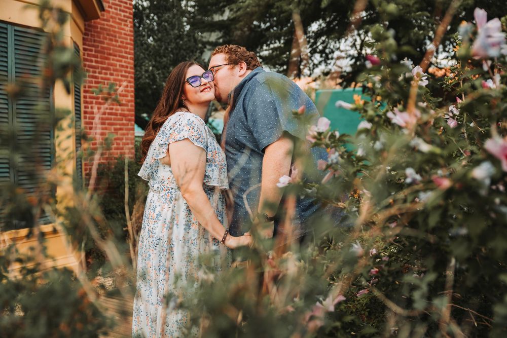 A couple shares a tender kiss in front of a brick building with green foliage framing the romantic moment.