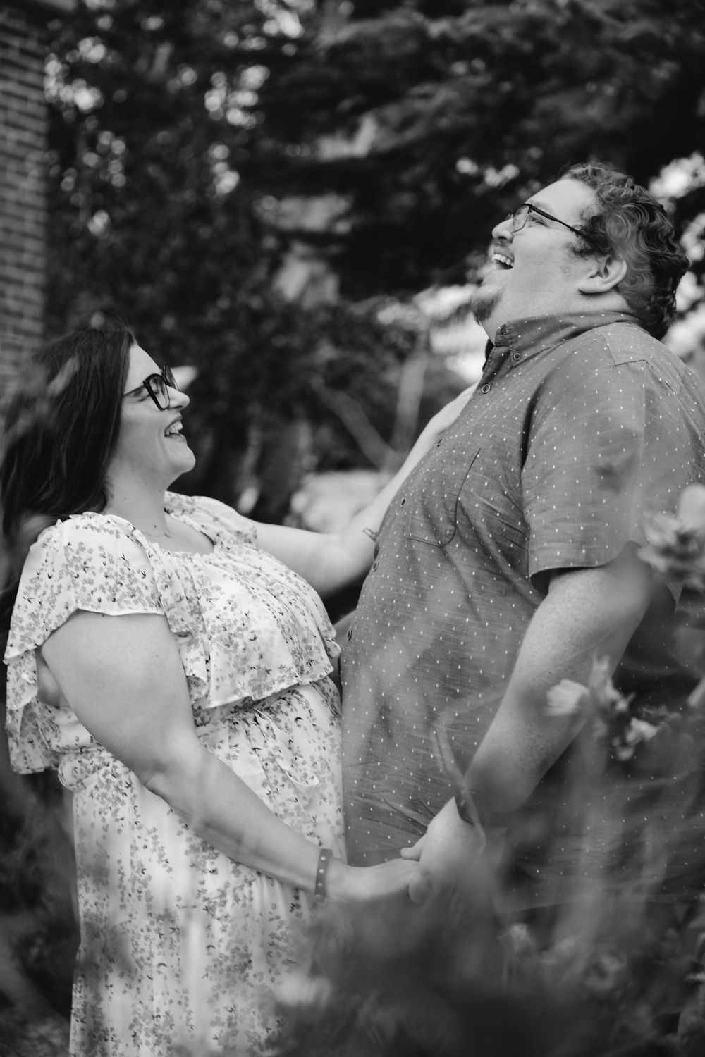 A couple shares a tender moment in black and white as they laugh together outdoors surrounded by greenery.