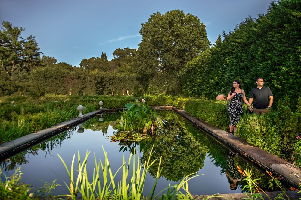 Couple strolling along a reflecting pool bordered by lush gardens and trees on a summer evening.
