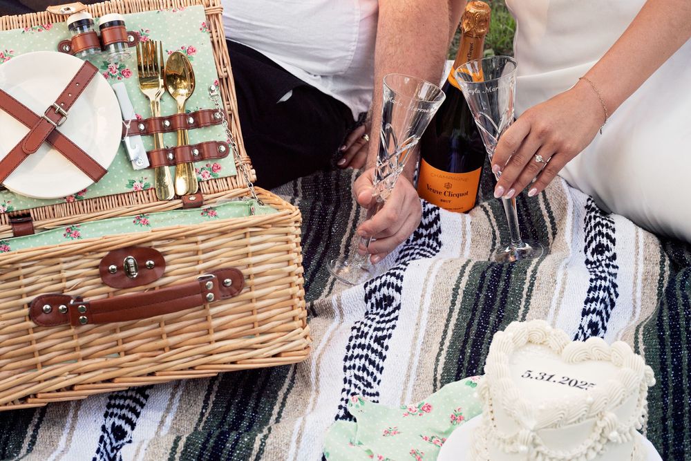 Hands pouring red wine into glasses at an outdoor picnic with a wicker basket and striped blanket in the background.
