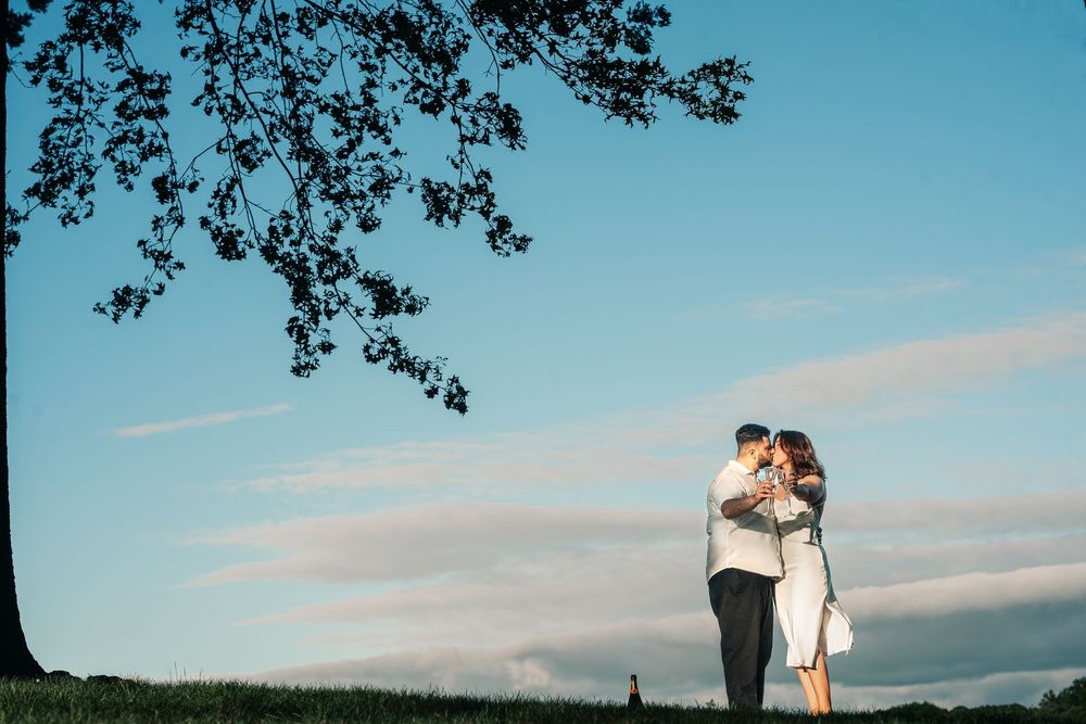 A couple in wedding attire stands silhouetted against a blue sky with tree branches framing the scene.