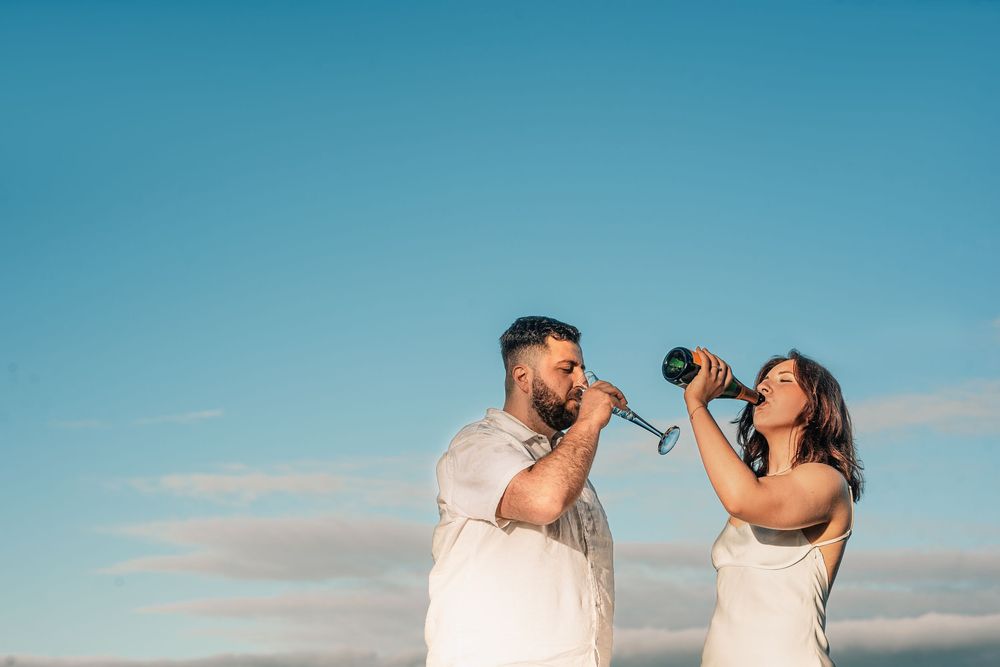 A couple in white outfits shares a romantic kiss on a beach with blue sky and ocean in the background.