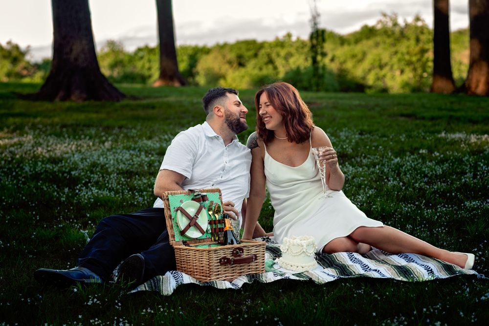 A romantic couple enjoys a summer picnic on a blanket in a lush green park at sunset.
