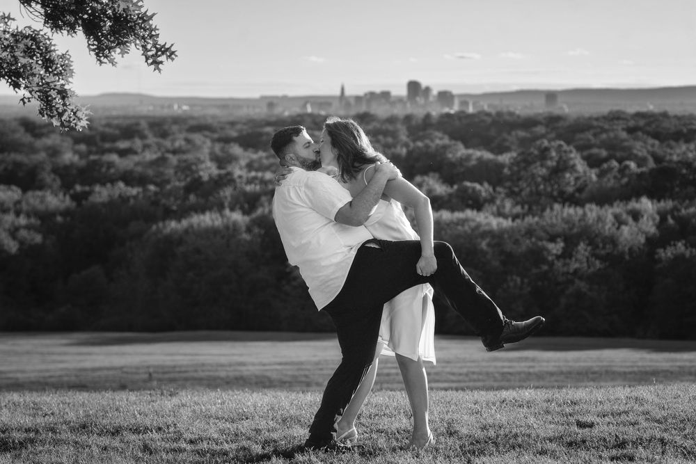 A romantic couple dances together in an outdoor field with rolling hills and trees in the background.