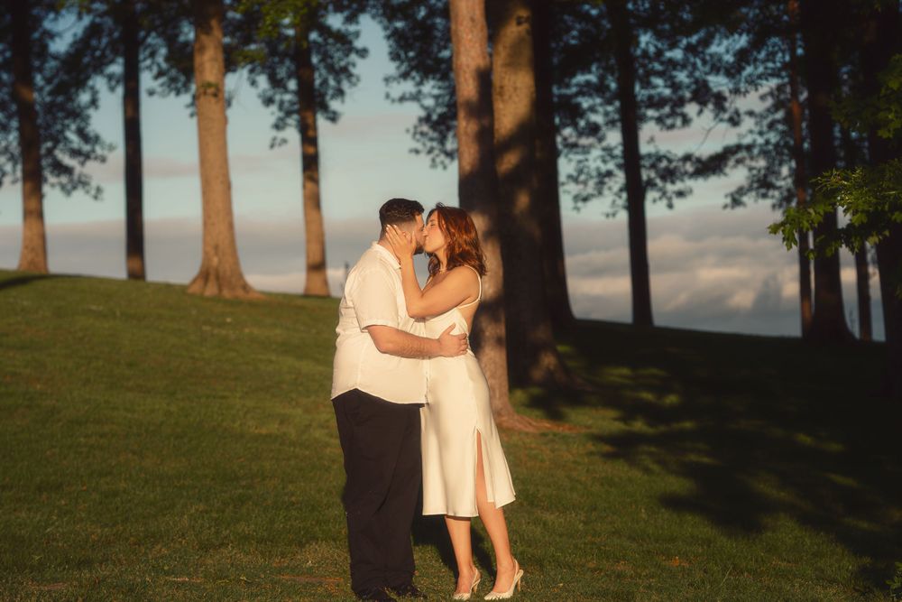 A romantic couple in white outfits embrace during a sunset photo session on a grassy hillside with trees in the background.