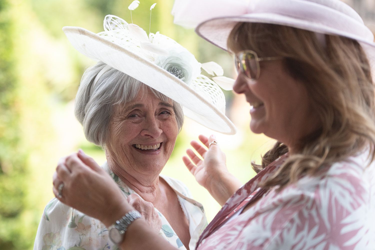 Two elegantly dressed women in fancy hats share a heartfelt laugh at an outdoor event.