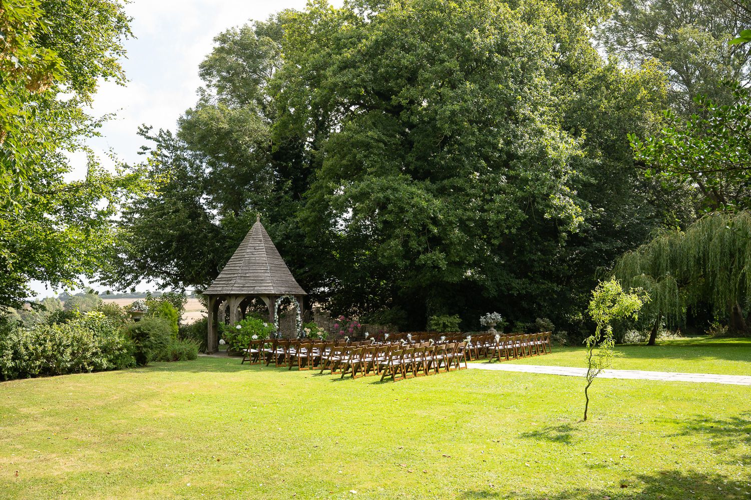 A panoramic view of a lush green outdoor wedding venue with wooden chairs set up on rolling hills and trees.