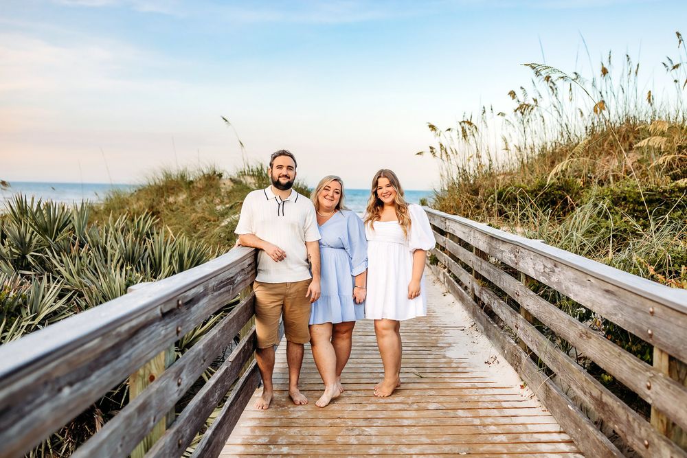 Three people walking on a wooden boardwalk path through beach dunes at sunset.