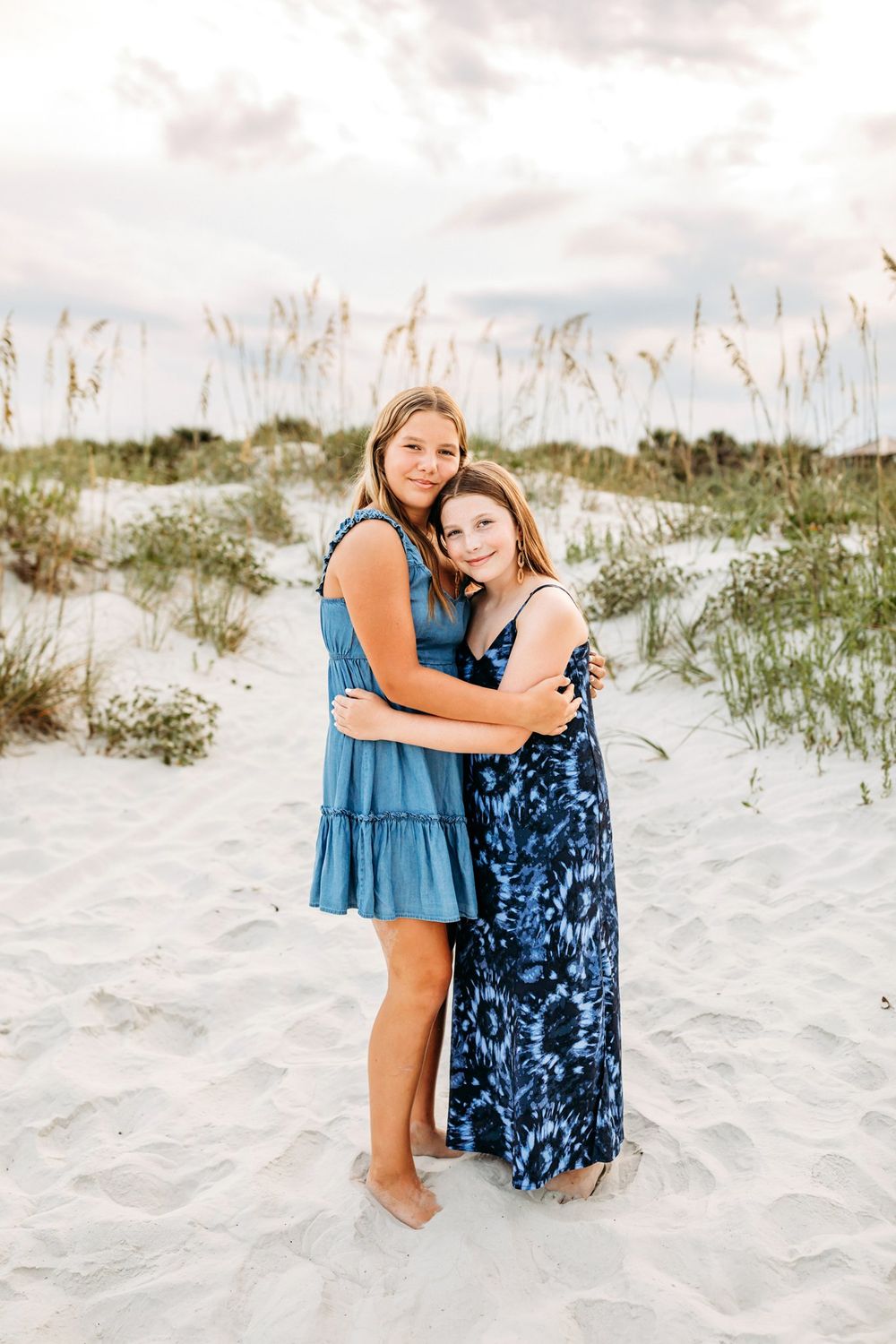 Two people in blue dresses sit together on a sandy beach with grass-covered dunes in the background.