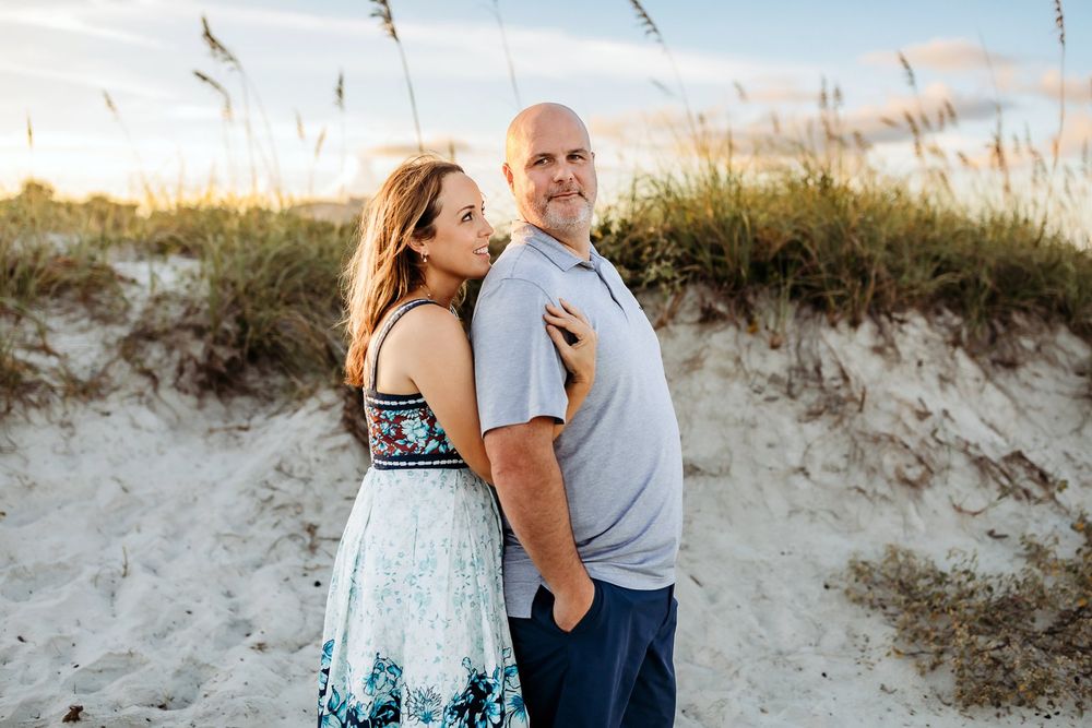 A couple embraces on a sandy beach path surrounded by sea oats and dunes during sunset.