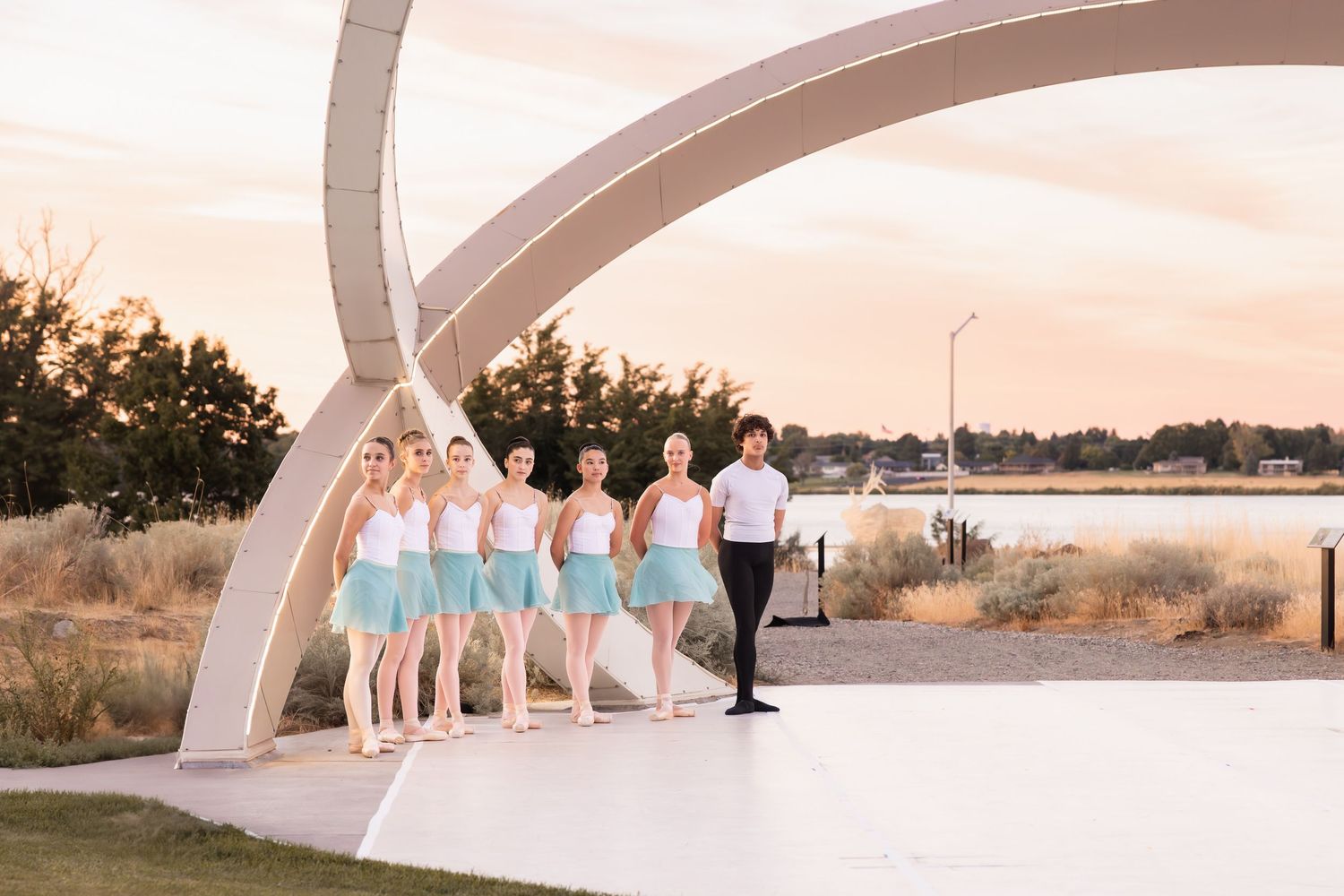 A wedding party poses under a modern white architectural arch at sunset by a waterfront.