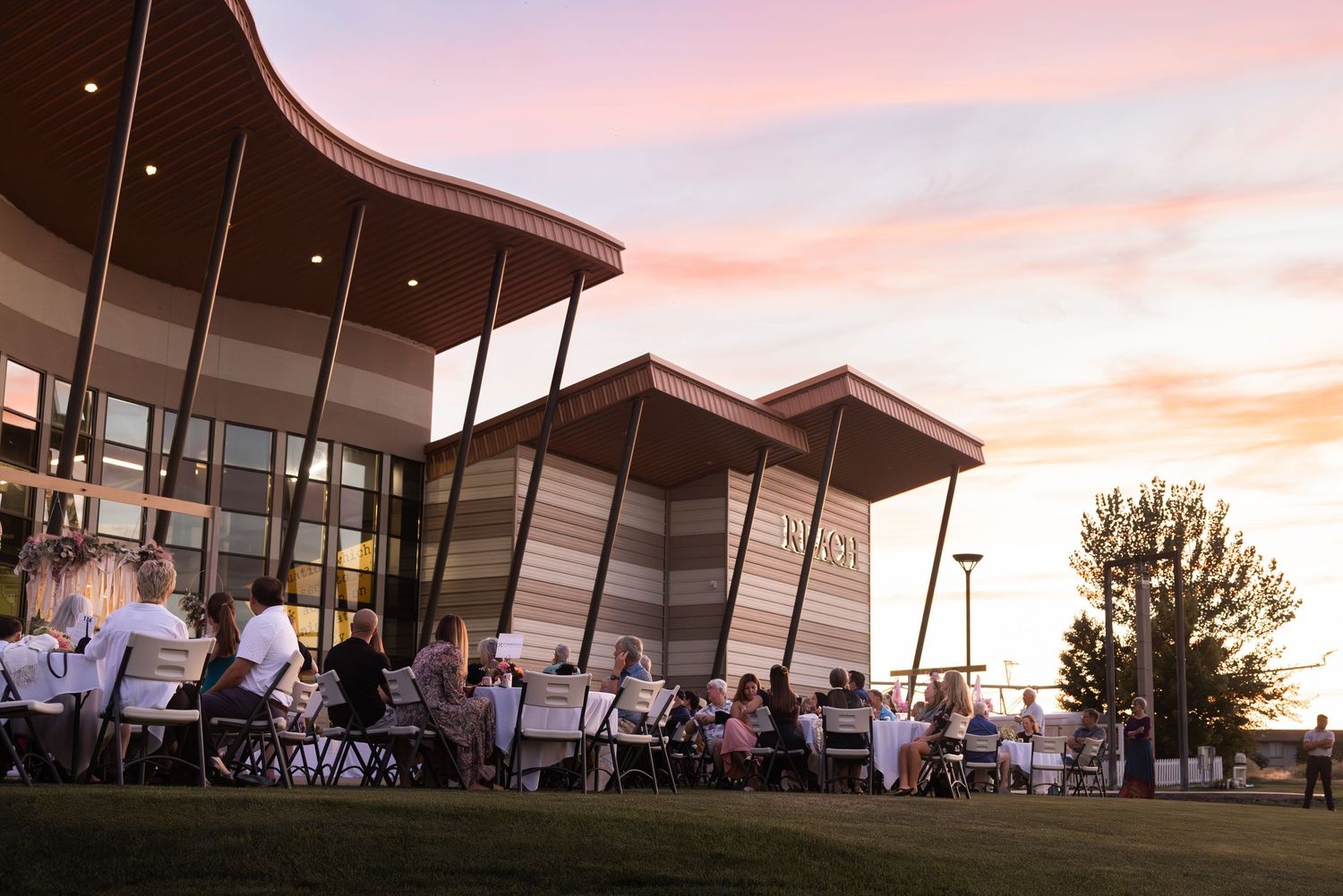 Modern outdoor event space with white canopy tents and seating arrangements glowing under a pink and orange sunset sky.