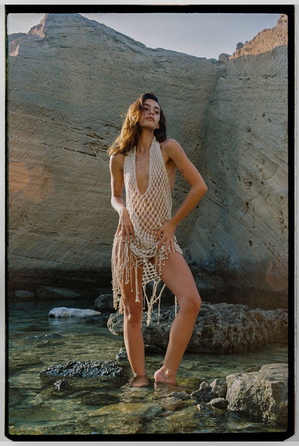 Figure posing in white crochet swimwear against rocky coastal backdrop during golden hour.