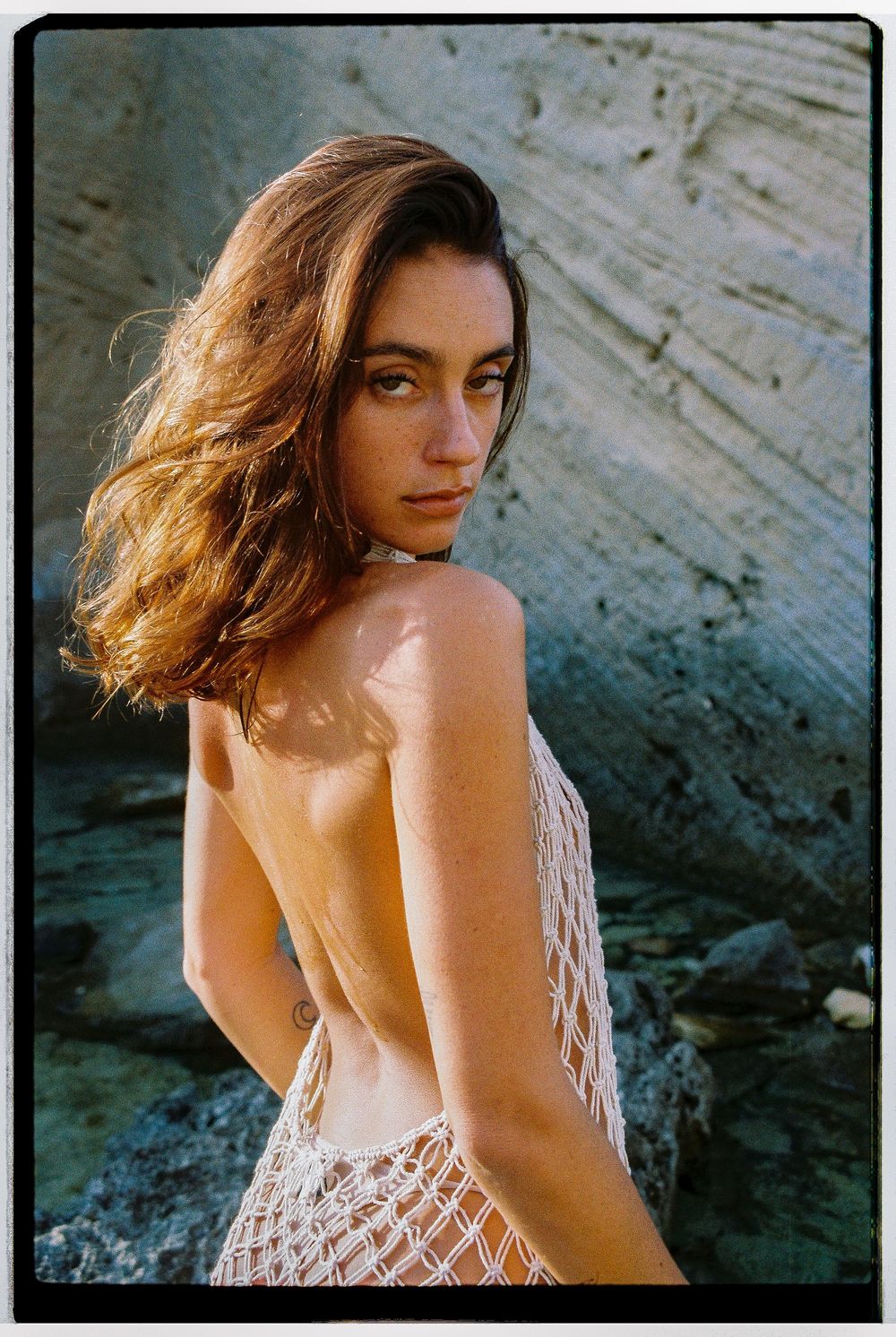 A woman in a backless crochet dress poses against a rocky backdrop in warm golden lighting.