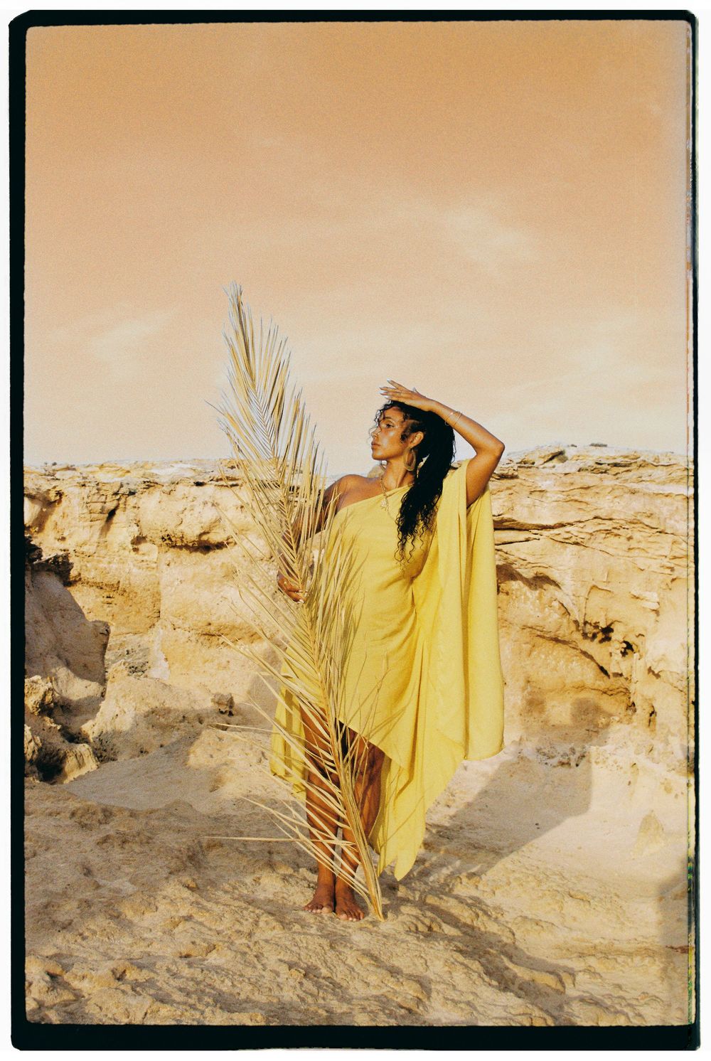 A figure in flowing yellow dress poses with palm frond against desert rock formations at dusk.