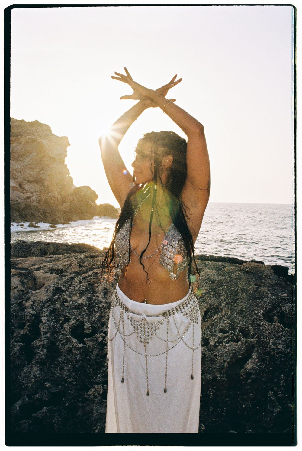 A dancer in white skirt and body paint poses gracefully against a rocky coastline at sunset.