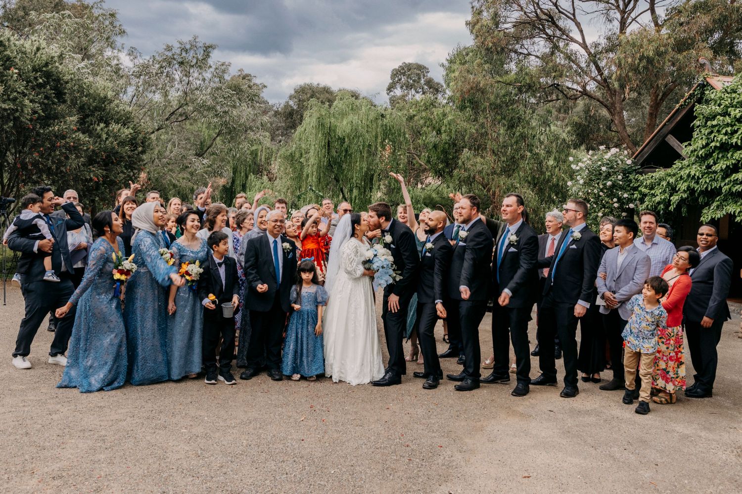 A panoramic wedding party group photo captured outdoors with guests lined up against a natural backdrop.
