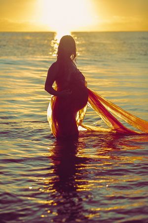 Silhouette in water at sunset with flowing fabric creating ripples on ocean surface.