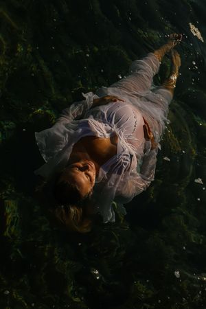 A figure in a white flowing dress lies ethereally in dark water surrounded by foliage and shadows.