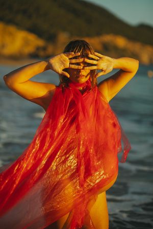 Figure in flowing red fabric stands in water during golden hour creating luminous effect.