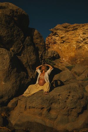 Person in white clothing posing dramatically on rocky coastal formations at golden hour.