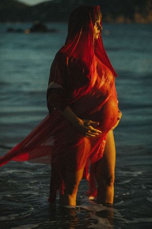A figure in flowing red fabric stands in shallow ocean water at sunset creating a dramatic silhouette.
