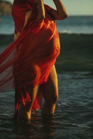 Red fabric flowing in water at sunset creates ethereal effect against dark beach.