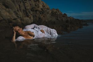 A figure in white fabric lies gracefully on coastal rocks during golden hour at the beach.