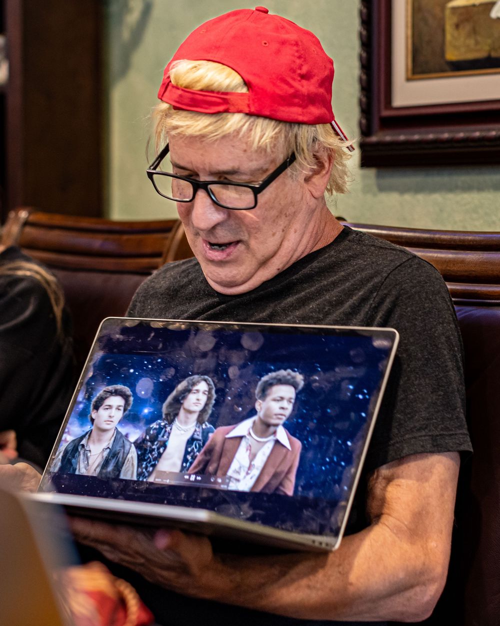 Someone in a red cap and glasses looks down at a tablet displaying a blue-tinted photo of three people.