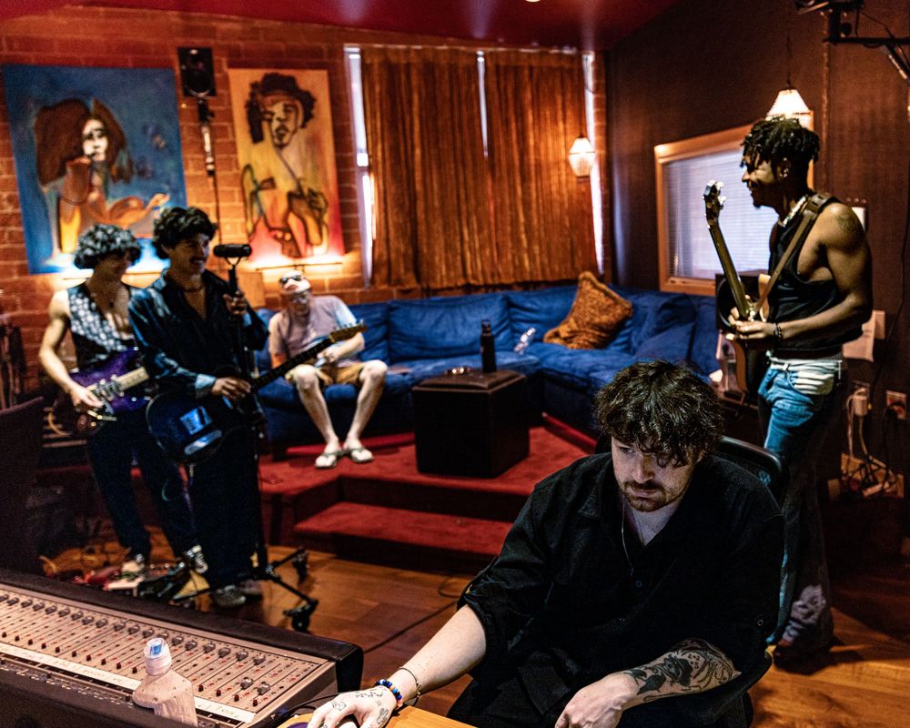 Recording studio session showing mixing console and musicians working in warm-lit wooden room.