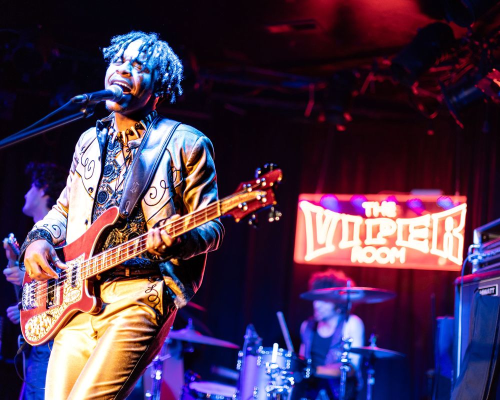 Musicians perform on stage at a dimly lit concert venue with red and blue lighting effects.