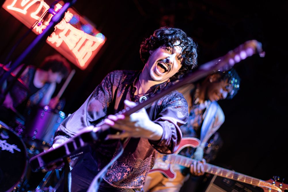 Musicians perform energetically on stage with guitars and instruments under dramatic purple lighting at a live concert.