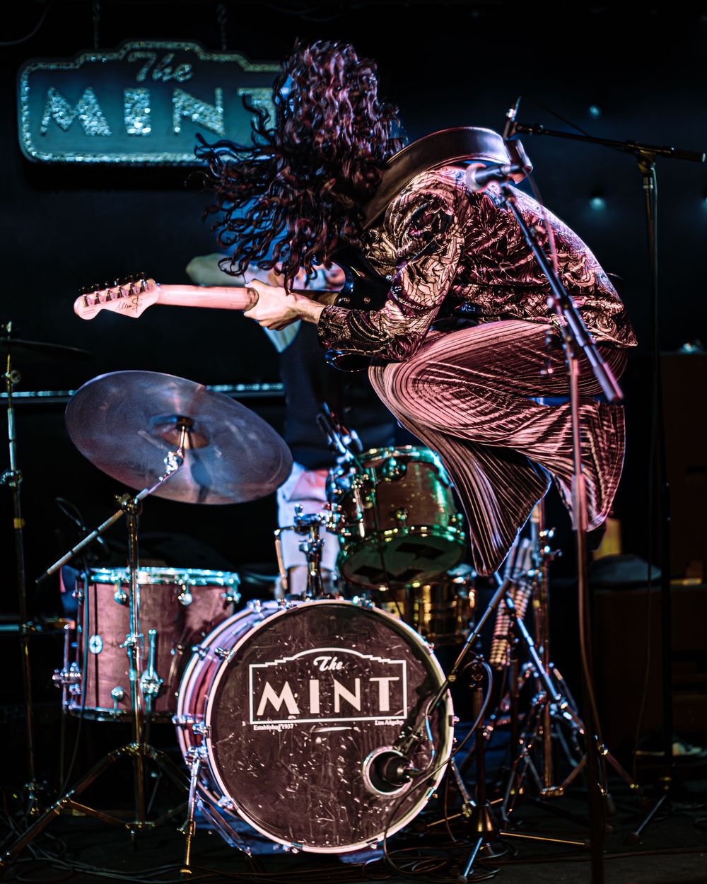 Energetic performer leaps high on stage with a guitar while performing at The Mint venue with a drum kit visible.