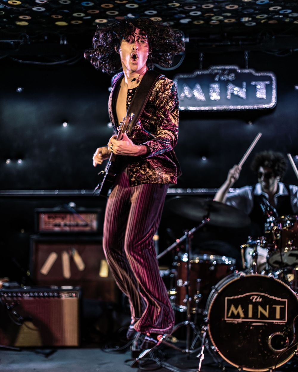 A guitarist performs on stage in a striped outfit under dramatic lighting at The Mint music venue.