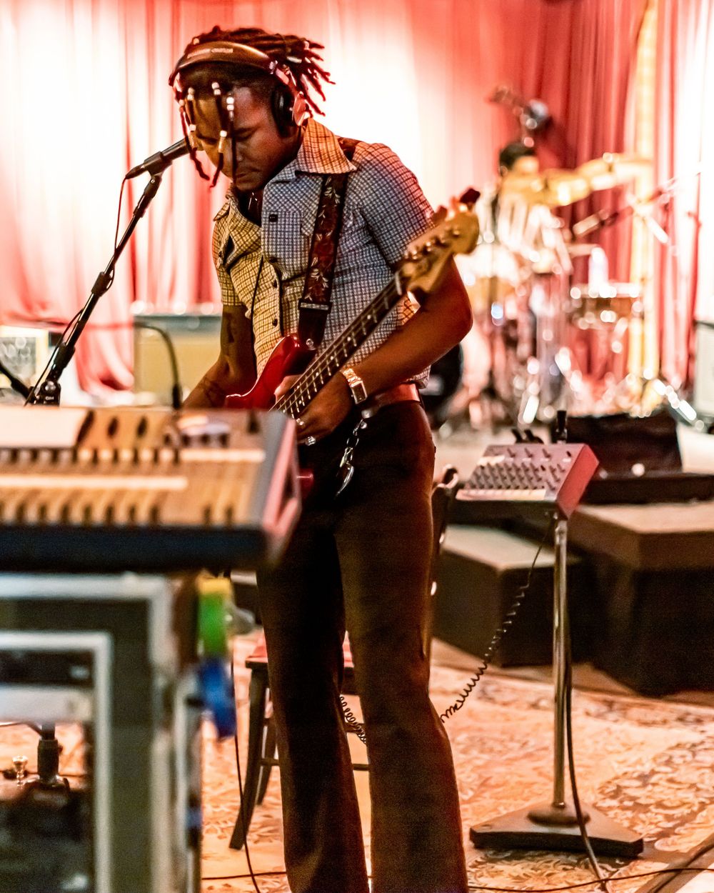 Musicians perform on stage with guitars and microphones in a warm-lit concert setting with red curtains in background.