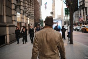 Person in brown coat walking down an urban street with tall buildings in the background.
