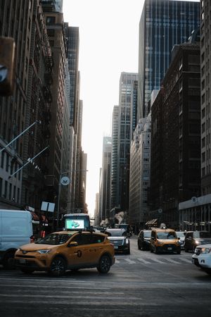 Yellow taxis line up on a busy Manhattan street between towering skyscrapers.