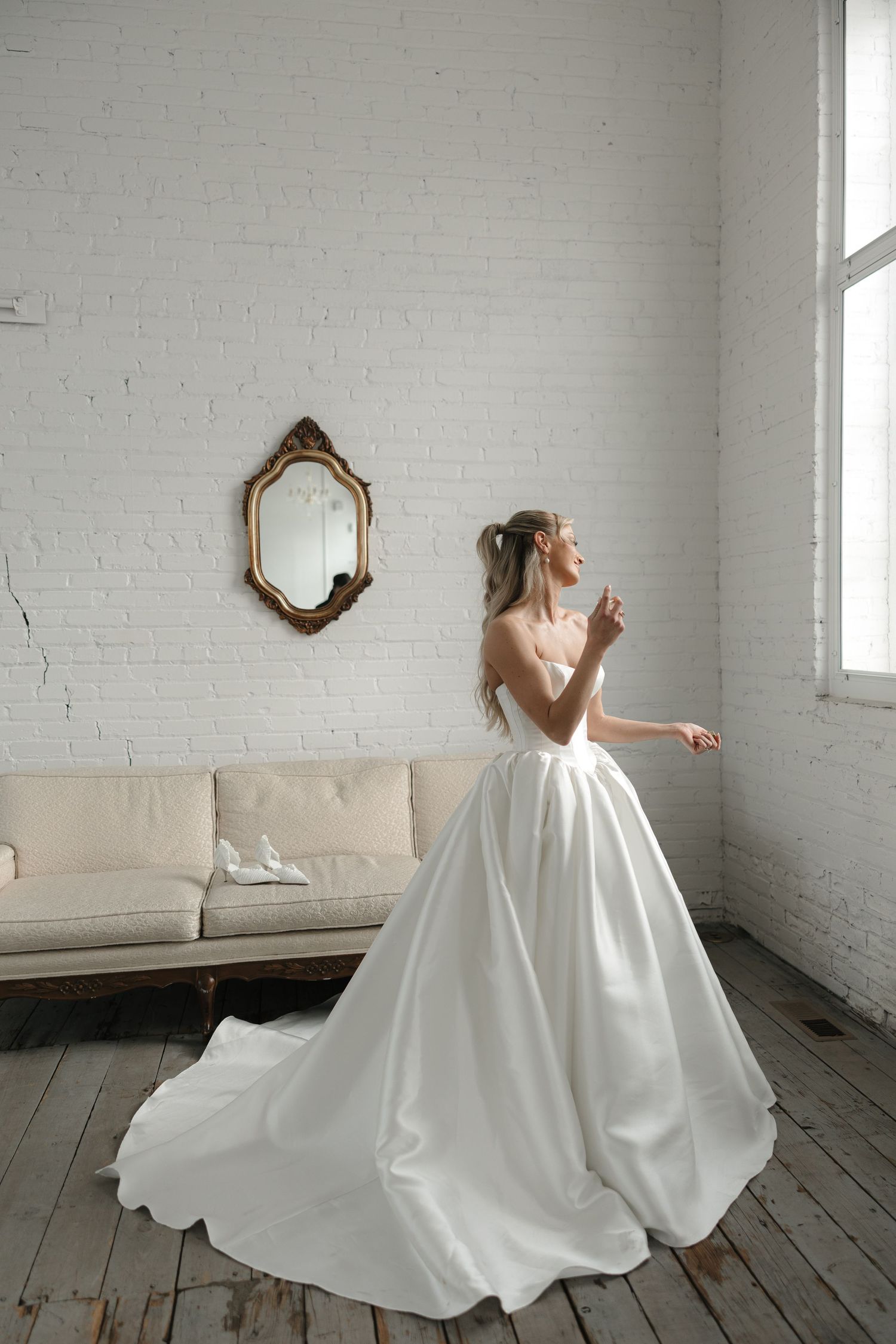 A flowing white wedding gown twirls gracefully across a rustic wooden floor in a bright minimalist room with white brick walls.